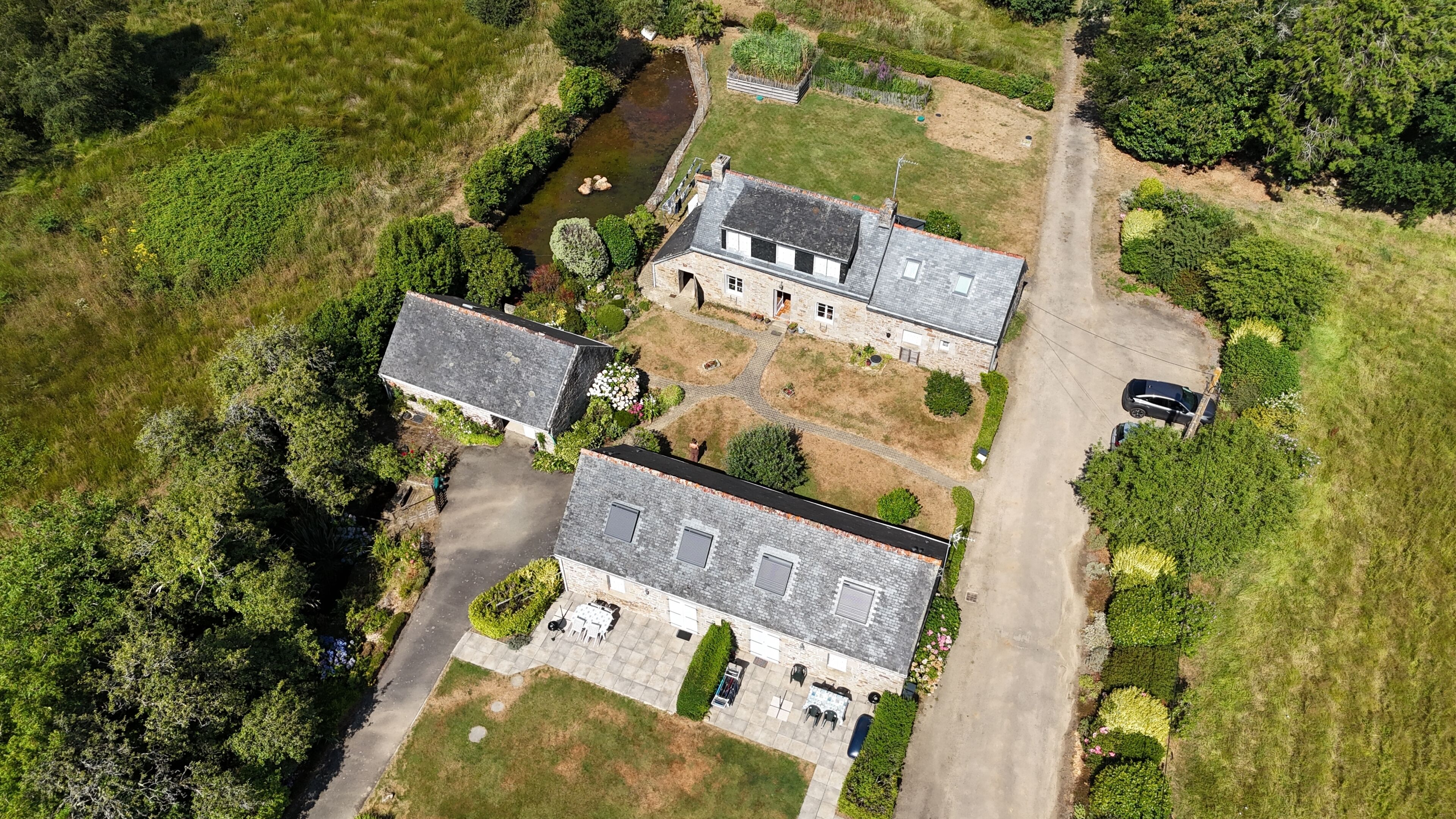 Vue aérienne d’une propriété comprenant une maison principale, un bâtiment plus petit, une allée et un ruisseau.