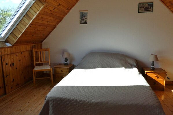 A bedroom with a sloped ceiling, a bed, two bedside tables with lamps, a wooden chair, and a small picture on the wall.