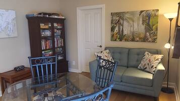 A living room with a glass-top dining table, two blue chairs, a blue sofa with patterned pillows, a bookshelf, a door, and a painting of tropical scenery.