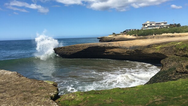 A coastal landscape with a rocky shoreline, waves crashing against the rocks, and a large building with multiple stories on a grassy hill.