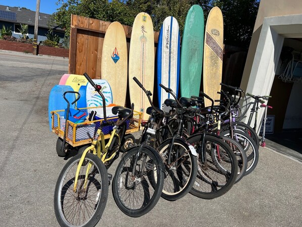 Varias tablas de surf apoyadas contra una pared, un carro de bicicleta con una caja azul, y varias bicicletas estacionadas al frente.