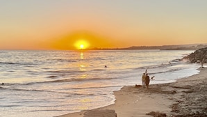Escena de playa con el sol poniéndose sobre el océano, olas suavemente acariciando la orilla, y algunas personas caminando por la playa.