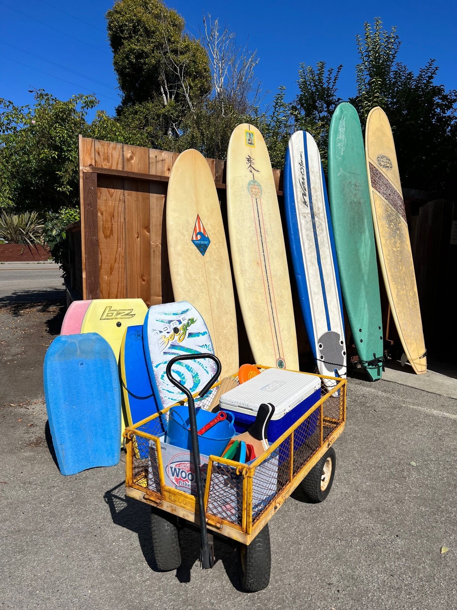 Ein Wagen mit Surfbrettern und einer Kühlbox, vor einem Holzzaun mit Bäumen im Hintergrund abgestellt.