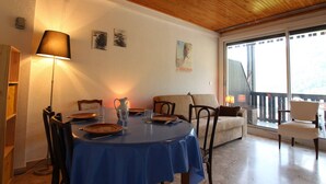 A dining area with a round table covered in a blue tablecloth, surrounded by wooden chairs. A lamp is on the left side, and there are two framed pictures on the wall. - Cozy Studio in Chantemerle - Sleeps 4, Parking, Balcony, 2 Stars (Saint-Chaffrey)
