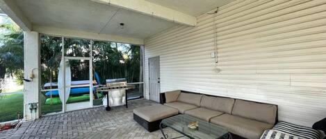 A covered patio area with a herringbone tile floor, a glass-top table, a beige sectional sofa, and a striped chair.