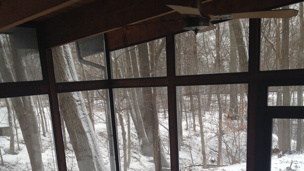 Ein Zimmer mit großen Fenstern und Blick auf einen verschneiten Wald. Zwei Holzstühle und ein grünes Sofa mit einer zusammengeklappten Decke sind vorhanden.