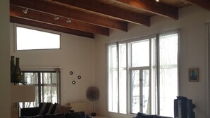 Ein Wohnzimmer mit einem Ecksofa, einem Couchtisch und einer Stehlampe. Große Fenster und eine Holzbalkendecke sind ebenfalls vorhanden.