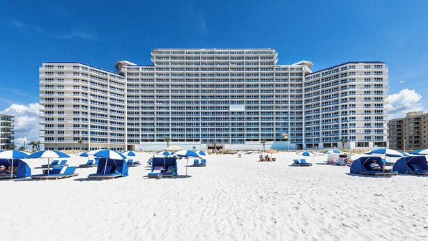A large hotel with multiple floors and balconies, a sandy beach in front with blue and white beach umbrellas, and lounge chairs. - West Corner w/Massive Balcony and Spectacular Gulf-Front Views! (Gulf Shores)