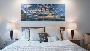 Un lit bien fait, avec une couette à rayures blanches et grises, deux lampes de chevet et un grand tableau représentant un paysage, placé au-dessus de la tête de lit.