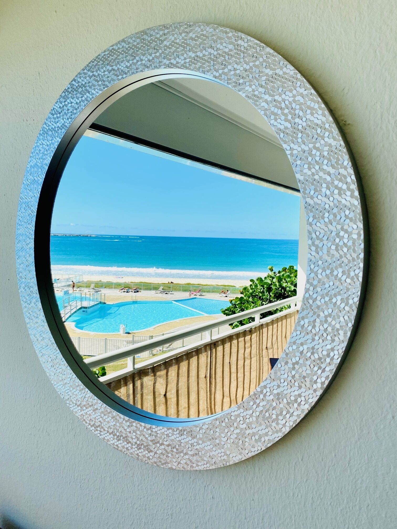 A round mirror with a textured frame reflecting a pool, a wooden deck, and a beach with the ocean in the background.