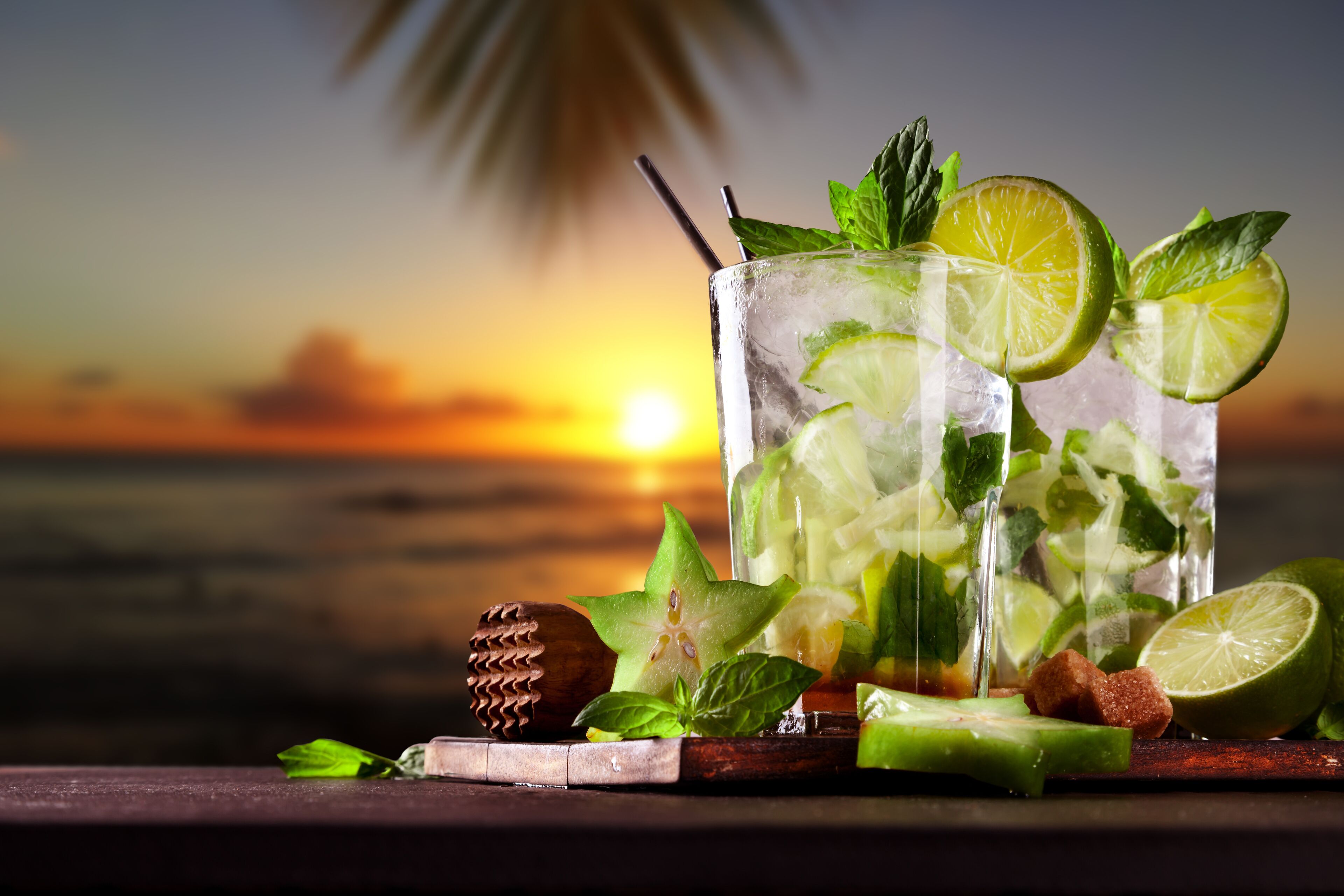 A glass of iced drink with lime and mint, placed on a wooden surface with a sunset background.
