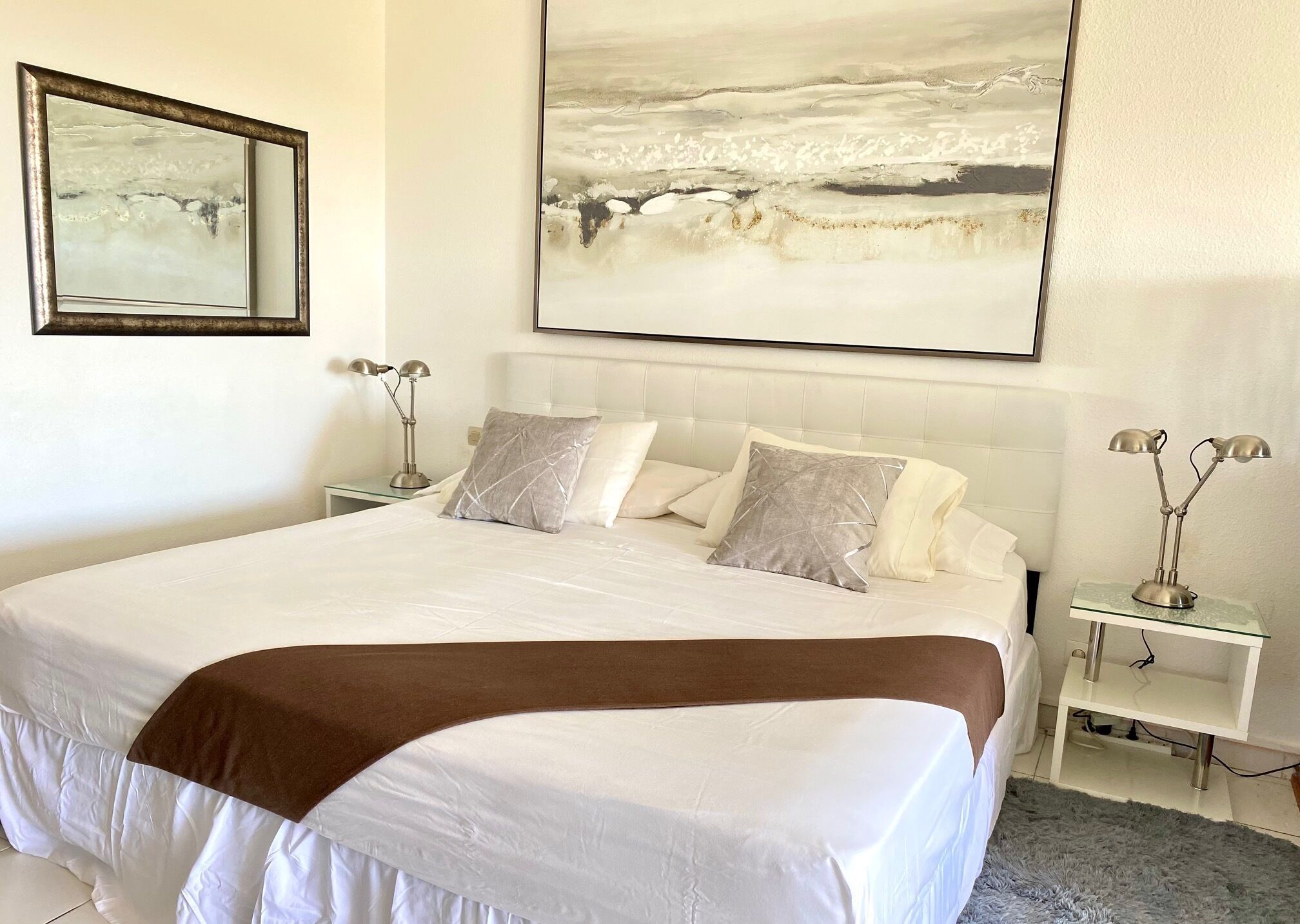 Een net opgemaakt bed met een wit-bruin sprei, twee bedlampen en een groot abstract schilderij erboven.