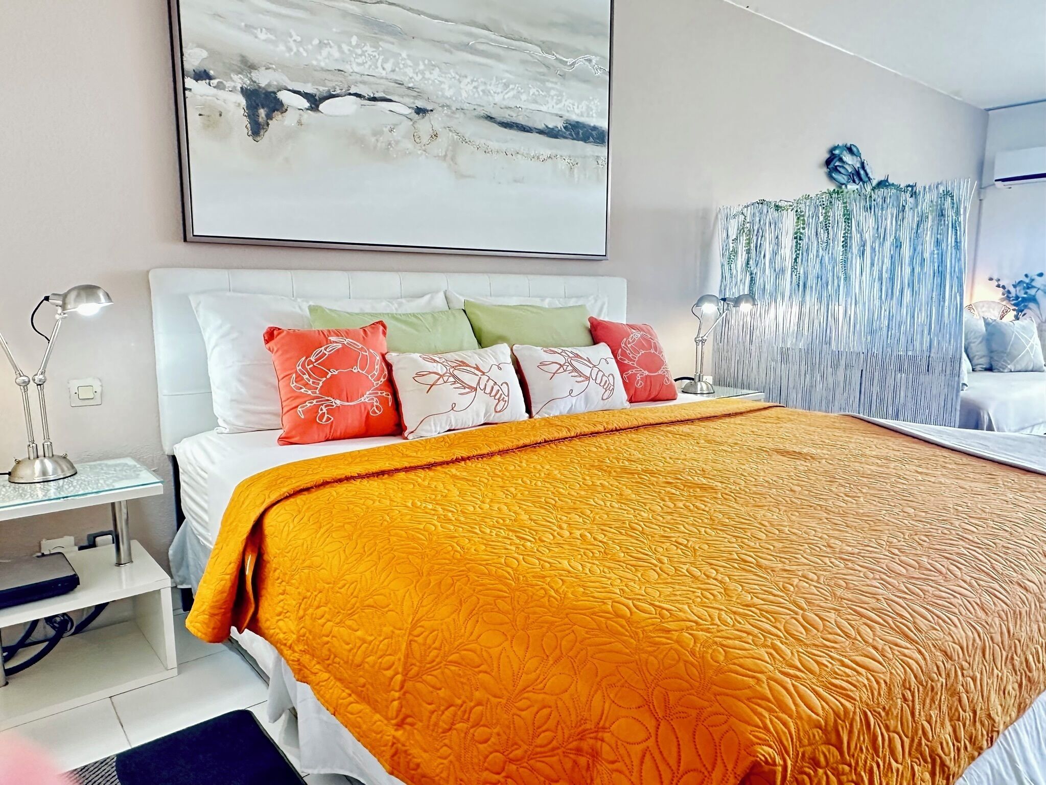 A bedroom with a large bed, white headboard, and multiple pillows. There is a nightstand with a lamp and a small table with a laptop. A large abstract painting is on the wall above the bed.