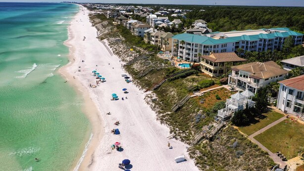Vue aérienne d’un hôtel situé en bord de mer, avec une longue plage de sable, des eaux turquoise et limpides, et plusieurs bâtiments surmontés de toits bleus.