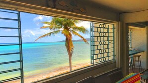 Una habitación con ventana que da a una playa y al océano, una palmera y un mantel a rayas de colores.