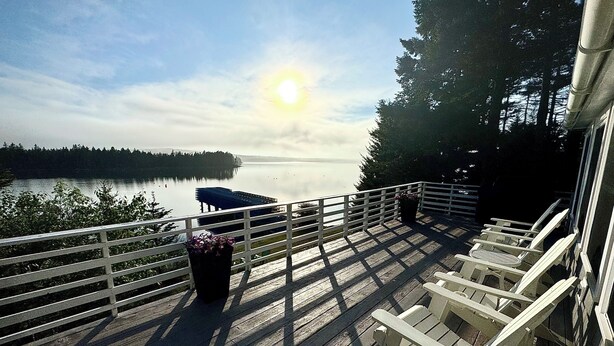 Une terrasse en bois avec des balustrades blanches, donnant sur un lac paisible où se trouve un quai, le tout entouré d’arbres.