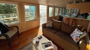 Un intérieur en bois comprenant un canapé, un fauteuil en osier, une petite table avec un livre et un coin cuisine avec un tabouret de bar.