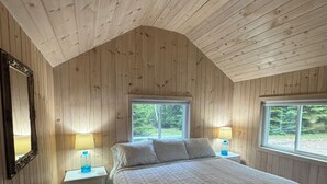 Une chambre avec un plafond en bois, un lit recouvert d’une couverture matelassée, deux tables de chevet avec des lampes et des fenêtres donnant sur un paysage verdoyant.