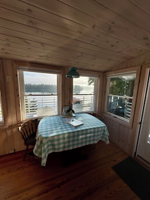 A room with a checkered table, a chair, a lamp, and large windows overlooking a lake.
