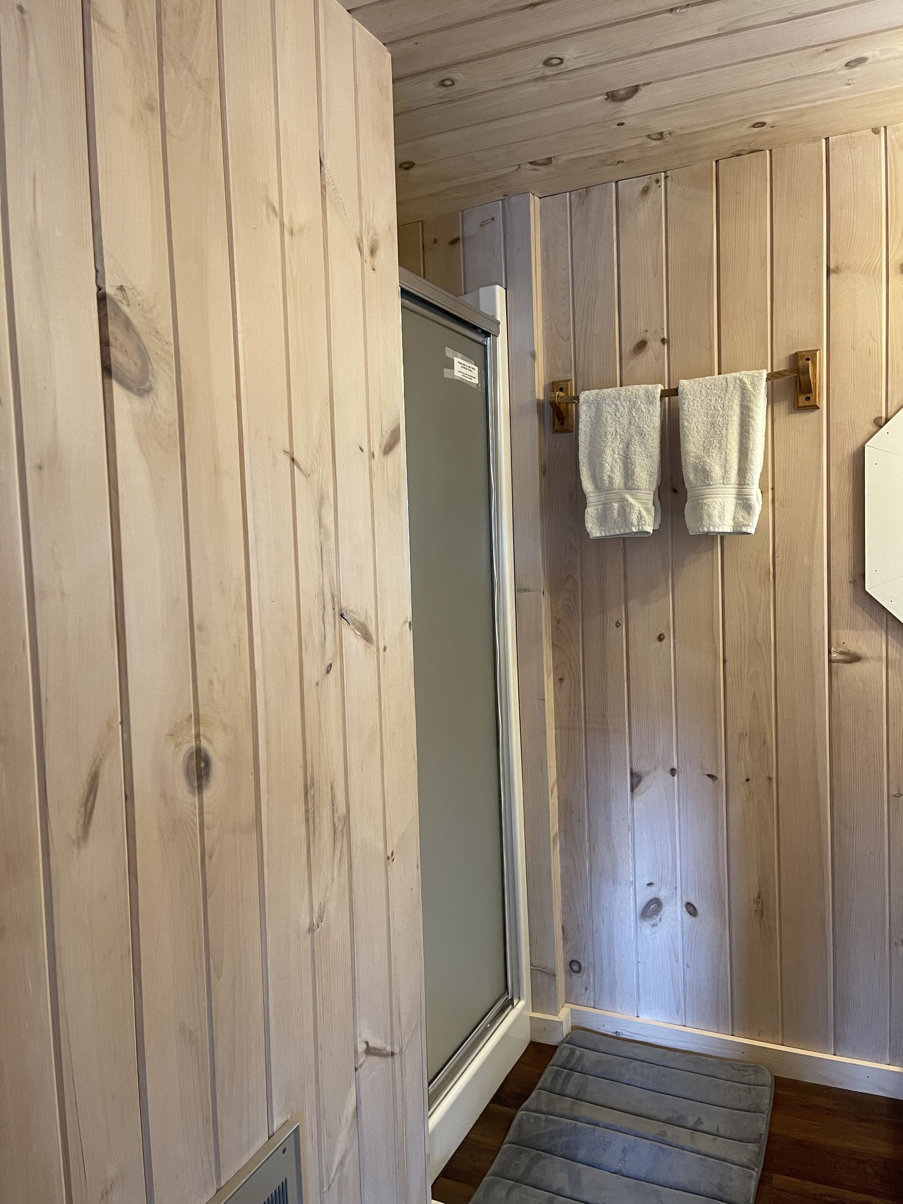 A wooden-paneled bathroom with a shower door, a towel rack with two towels, and a visible vent.