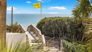 A wooden staircase leading to a beach with a flag, surrounded by greenery and a clear sky. - BEACHFRONT BUILDING l PRIVATE BEACH l GULF GLIMPSE l SEASONAL BEACH SETUP (Santa Rosa Beach)