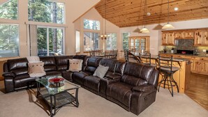 A spacious living room with a leather sectional sofa, a glass coffee table, and a dining area with a wooden table and chairs. The kitchen features wooden cabinets and a large island with bar stools. - S. Tahoe Luxury Home, 5 Minutes to Heavenly lodge (South Lake Tahoe)