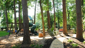 A playground with swings and a slide, surrounded by trees and a walking path.