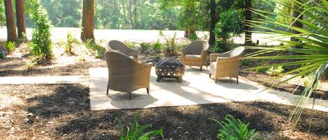Un patio amueblado con mobiliario de mimbre y una mesa central, rodeado de árboles y plantas.