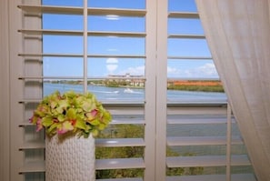 A window with horizontal blinds, a view of a body of water, and a white vase with flowers.