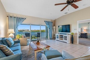 A spacious living area with a sofa, coffee table, and TV, a balcony with chairs, and a ceiling fan.