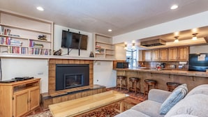 Una sala de estar con chimenea, un televisor montado en la pared y una zona de cocina con taburetes de bar.