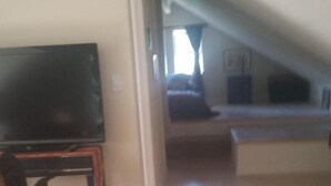 A living room with a flat-screen TV on a stand, a small table with magazines, and a view into an adjacent room with a bed and window.