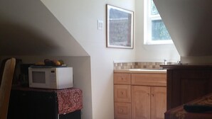 A small, dimly lit attic room with a kitchenette, a microwave, and a window.