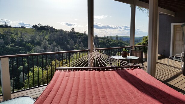 A balcony with a hammock, a table with a vase, and a view of a forested landscape.
