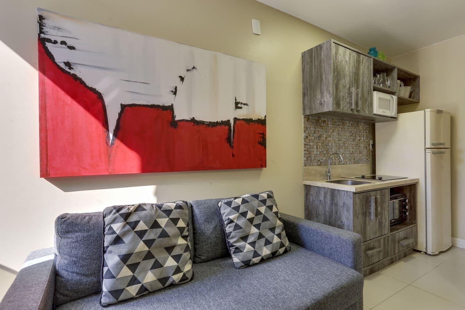 A living room with a grey sofa, two patterned cushions, a red and white abstract painting, and a kitchen with a wooden cabinet and a white refrigerator.