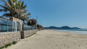 A beach with a stone wall and palm trees, a clear sky, and mountains in the background. - Pietra Flat (Florianópolis)