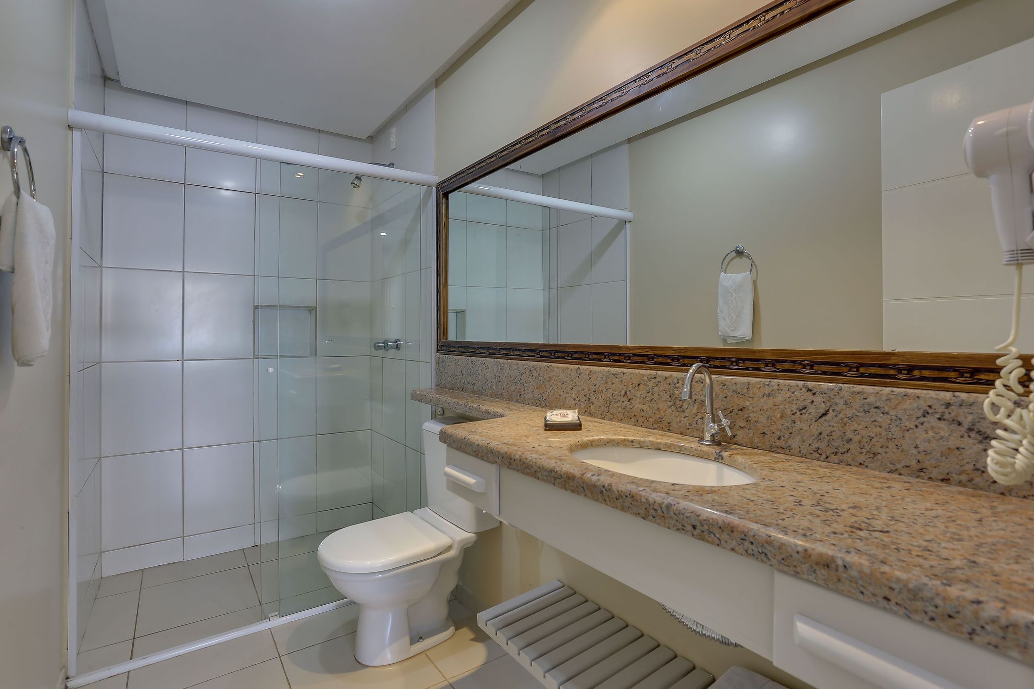 Un baño moderno con ducha envuelta en vidrio, inodoro blanco, lavamanos con encimera de granito y un espejo grande con marco de madera.
