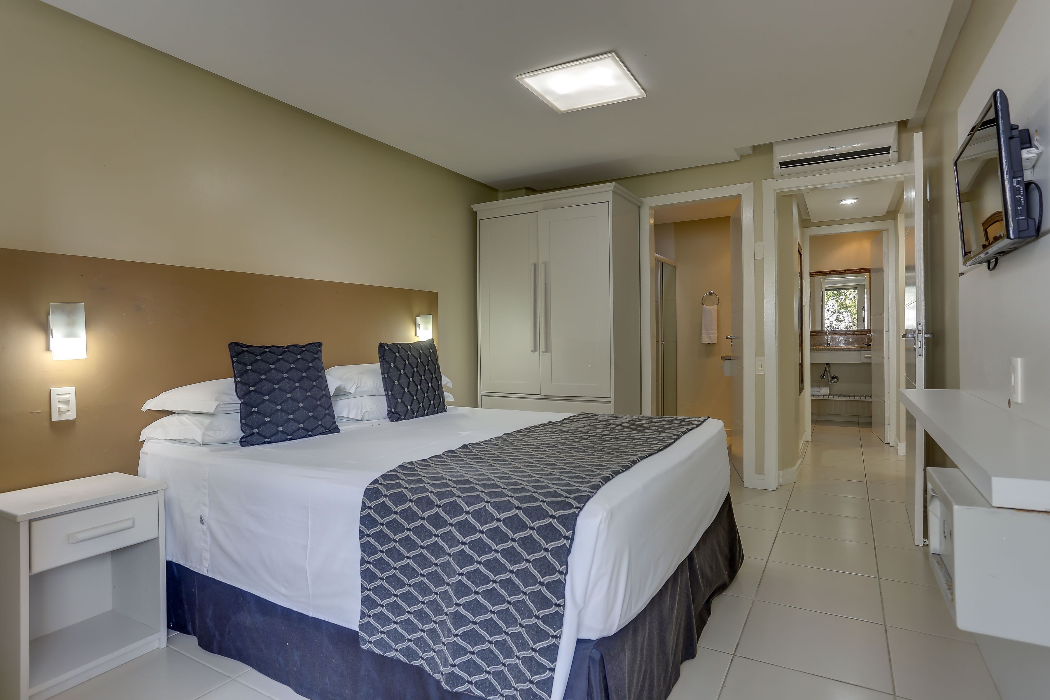 Un dormitorio con cama, mesitas de noche, un armario y un baño visible a través de una puerta abierta.