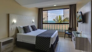 A hotel room with a bed, a desk, a chair, a TV, and a balcony with a view of the sea. - Pietra Flat (Florianópolis)