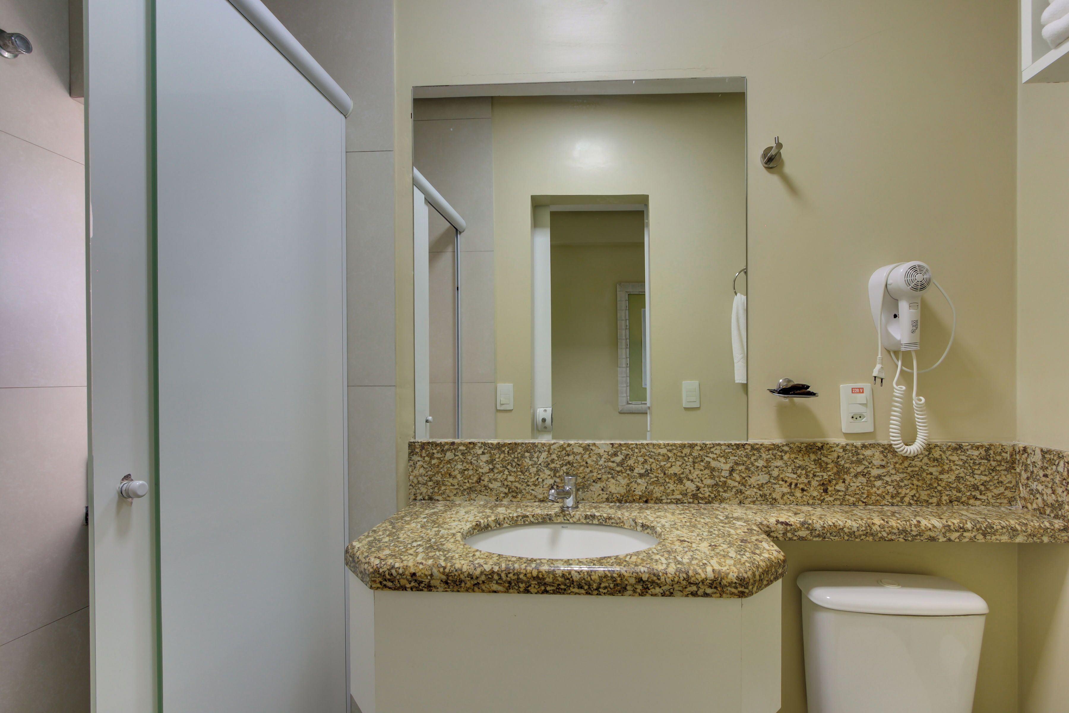 A bathroom with a granite countertop sink, a mirror, a hairdryer, and a toilet.