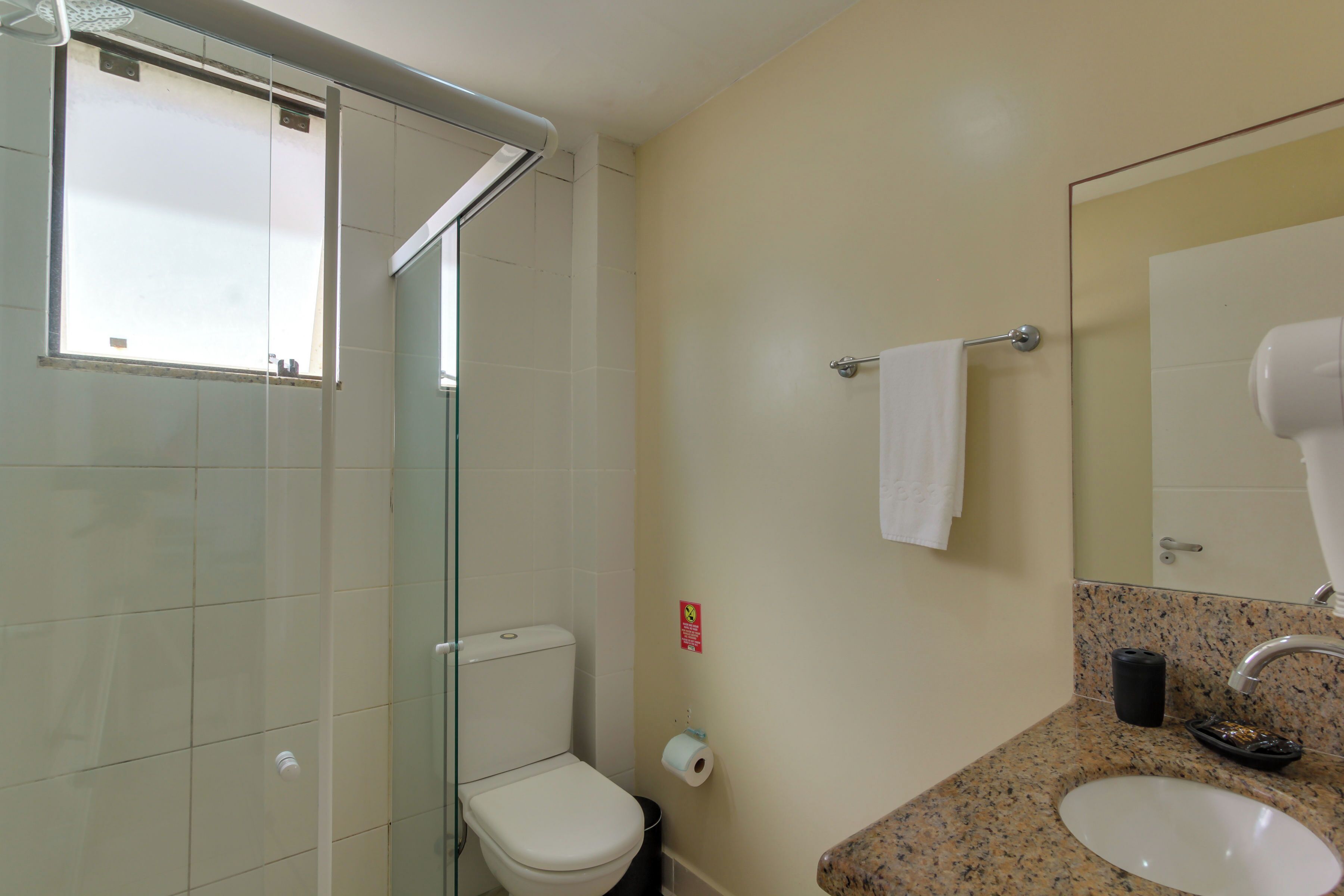 Et badeværelse med glasvæg omkring bruseren, toilet, håndvask med spejl og et håndklædestativ.