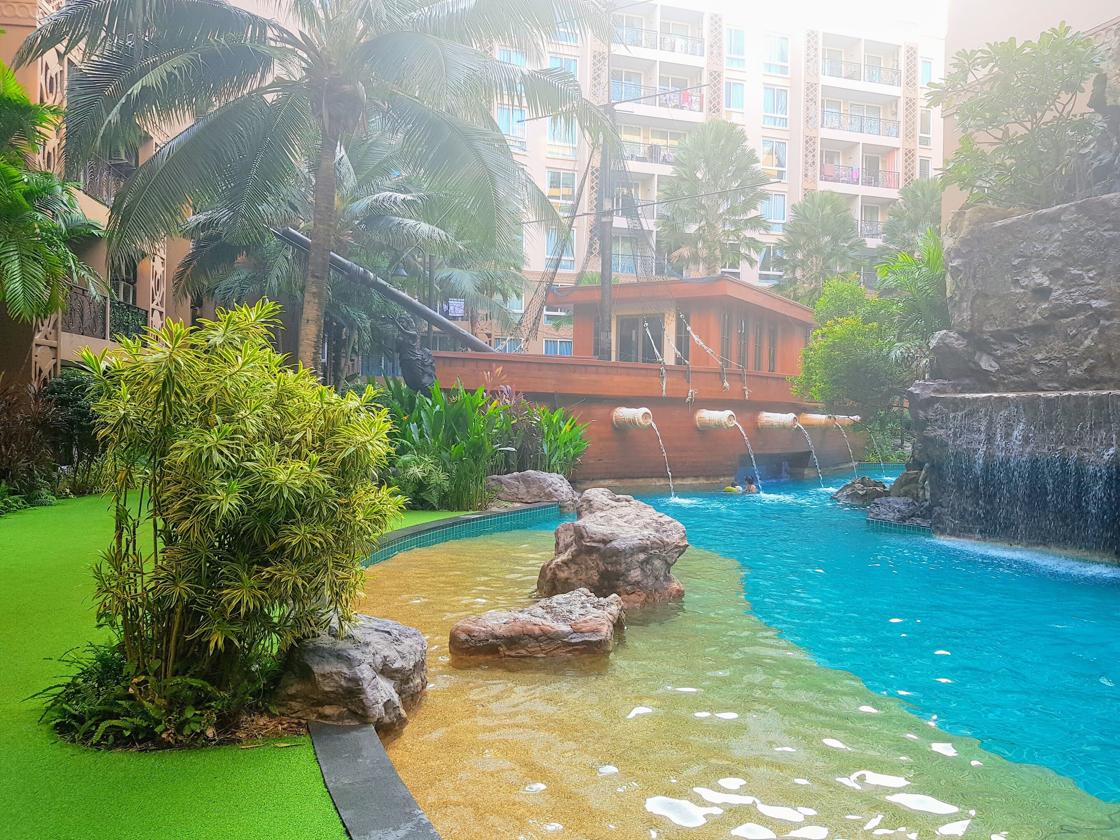 Uma área de piscina com água cristalina, rodeada por plantas tropicais e um edifício com varandas.