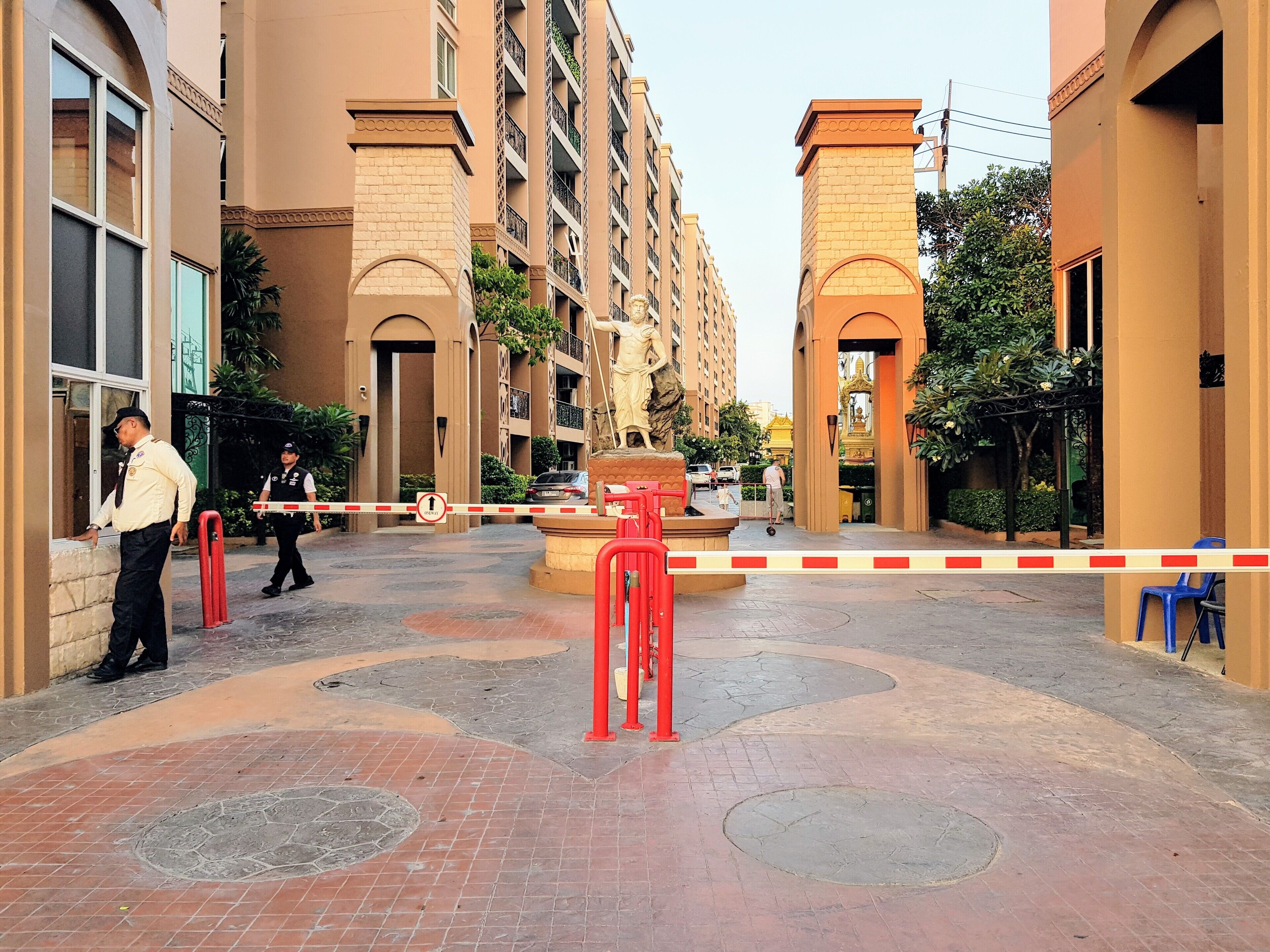 Une entrée sécurisée menant à un immeuble résidentiel, avec une barrière rouge, un homme en uniforme à l'entrée et une personne sur un scooter à l'arrière-plan.