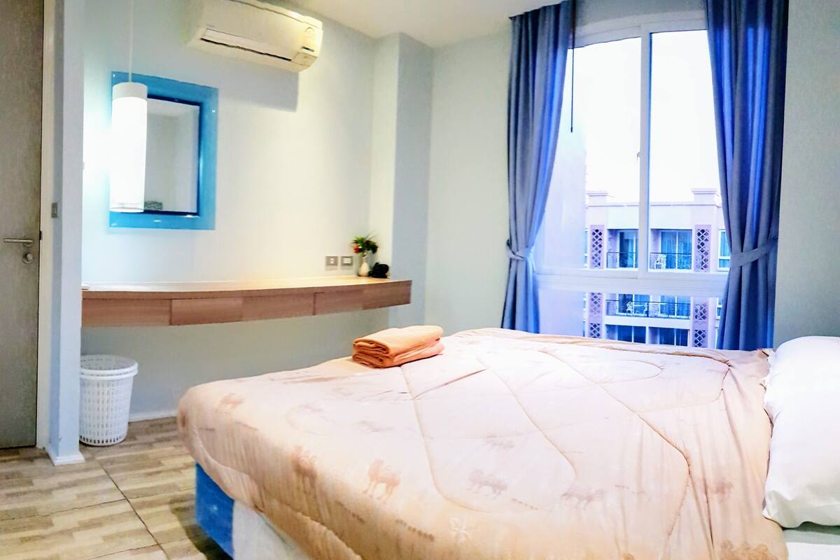 Une chambre d’hôtel avec un lit, une fenêtre avec des rideaux, un climatiseur fixé au mur, une petite étagère avec un vase et une porte.