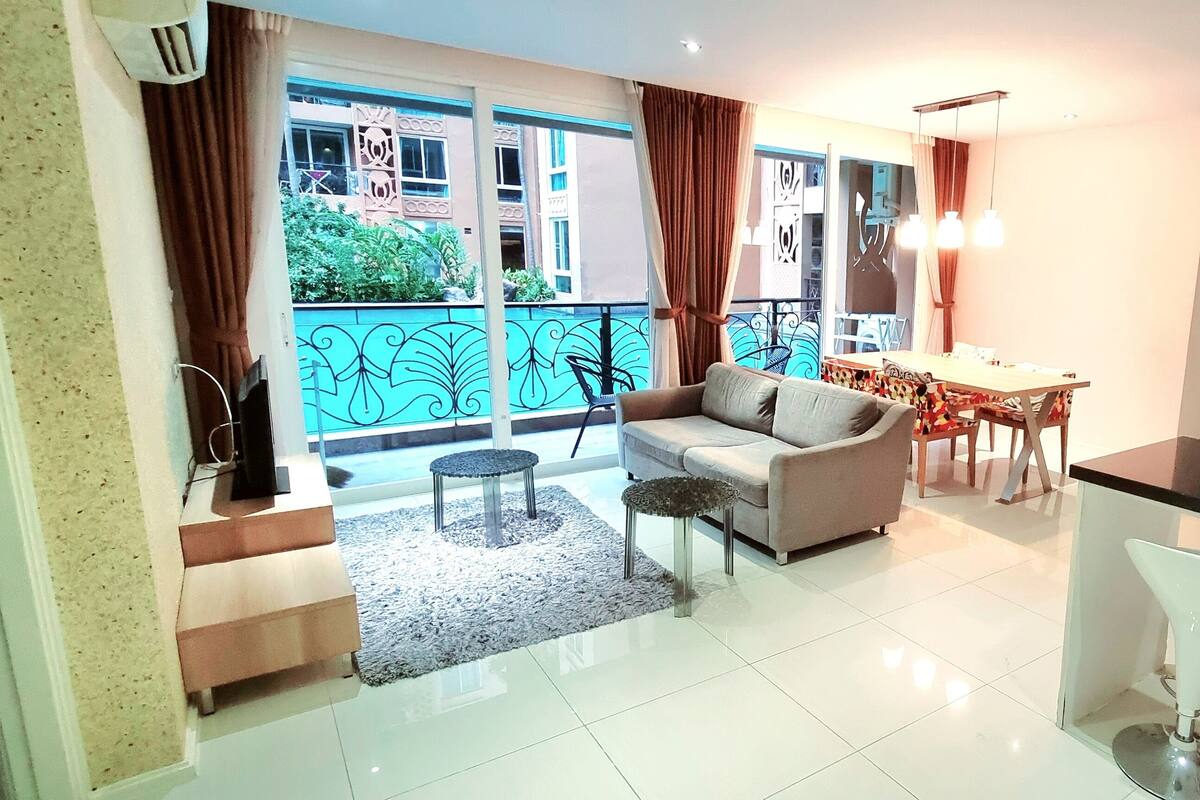 Un salon moderne avec un canapé, une table basse et un coin repas. Il y a un balcon avec vue sur les bâtiments et la verdure. La cuisine est dotée de meubles blancs et d’un plan de travail noir.