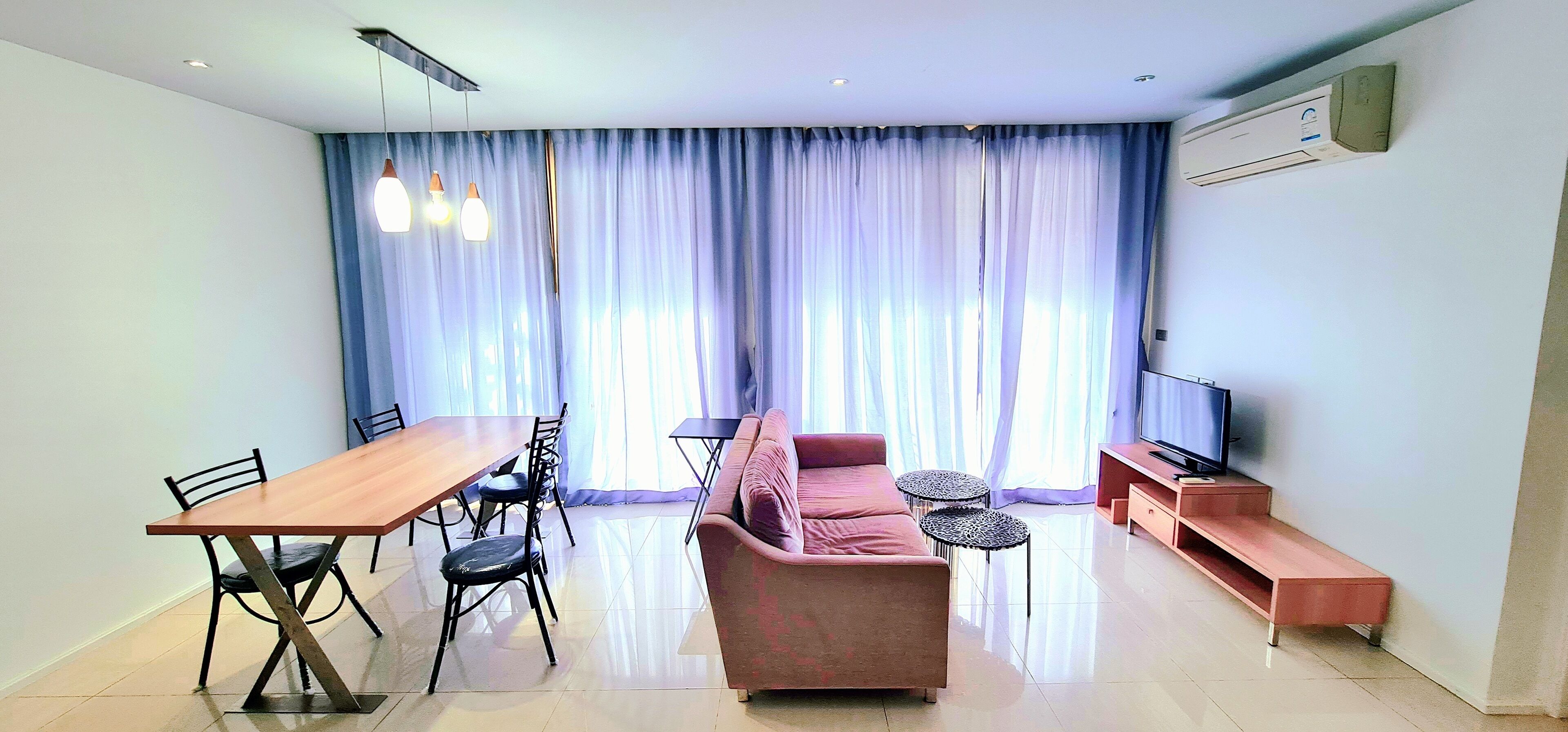Una sala de estar moderna con una mesa de comedor de madera, sillas negras, un sofá rosa, dos pequeñas mesas redondas, un televisor y un equipo de aire acondicionado en la pared.