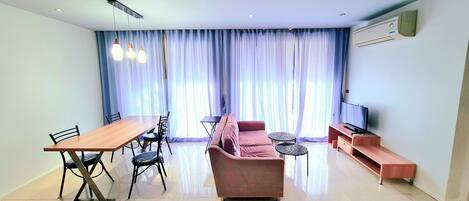 Una sala de estar moderna con una mesa de comedor de madera, sillas negras, un sofá rosa, dos pequeñas mesas redondas, un televisor y un equipo de aire acondicionado en la pared.