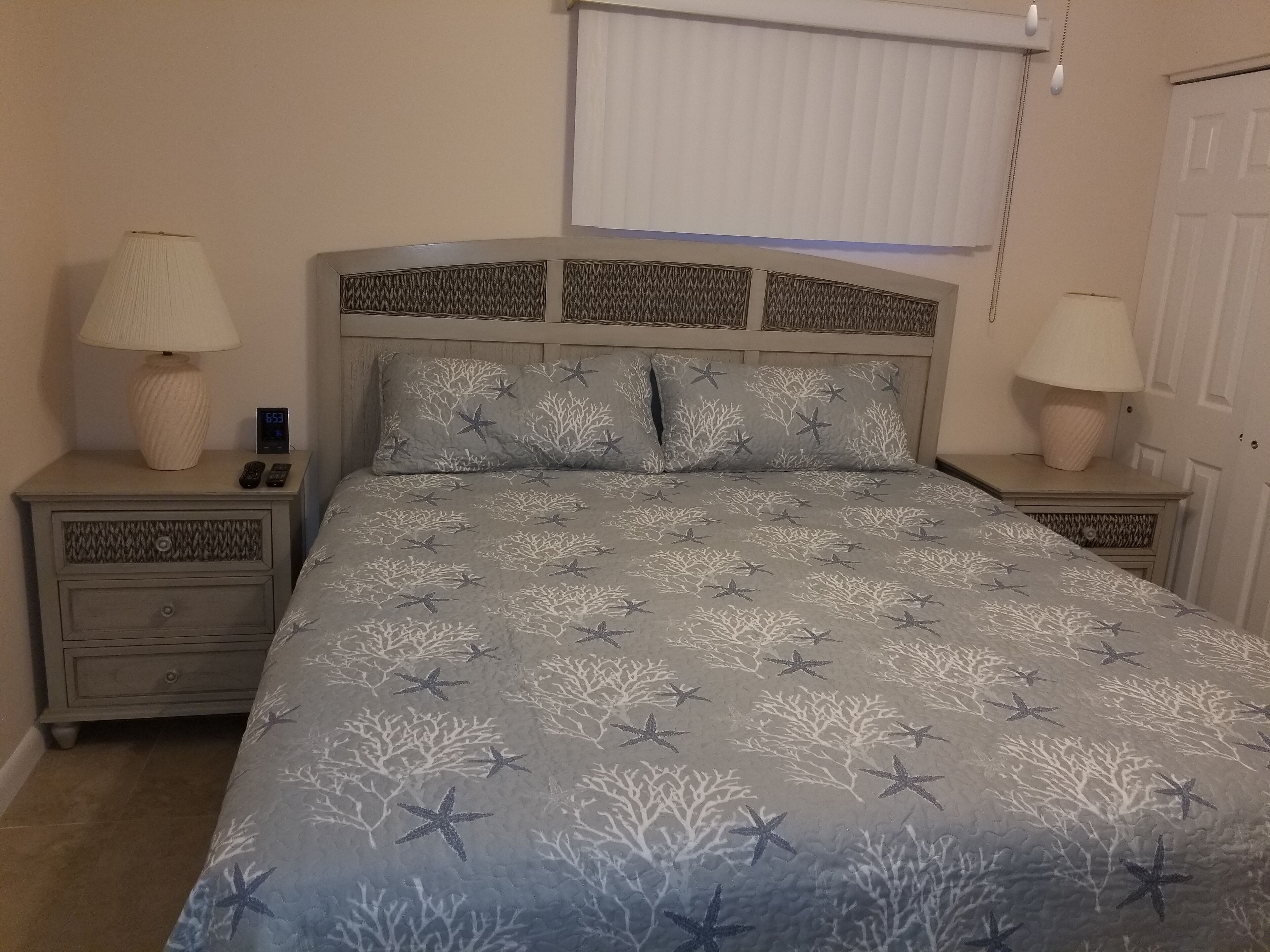 A bedroom with a bed featuring a starfish and coral pattern, two bedside tables with lamps, a nightstand with a clock, and a window with blinds.