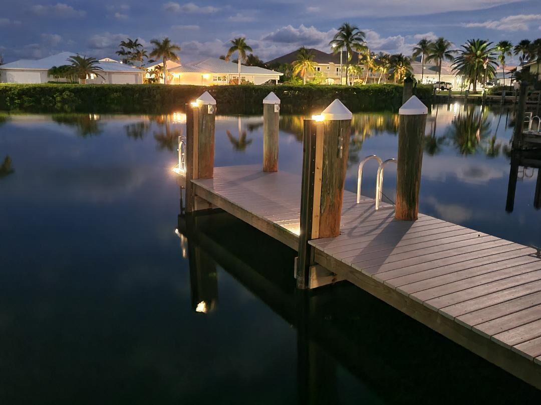A wooden dock extending into calm water with a reflection of palm trees and buildings.