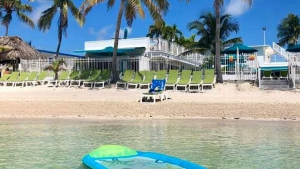 Ett hotell vid stranden med palmer, kristallklart blått vatten och ett poolområde med solstolar.